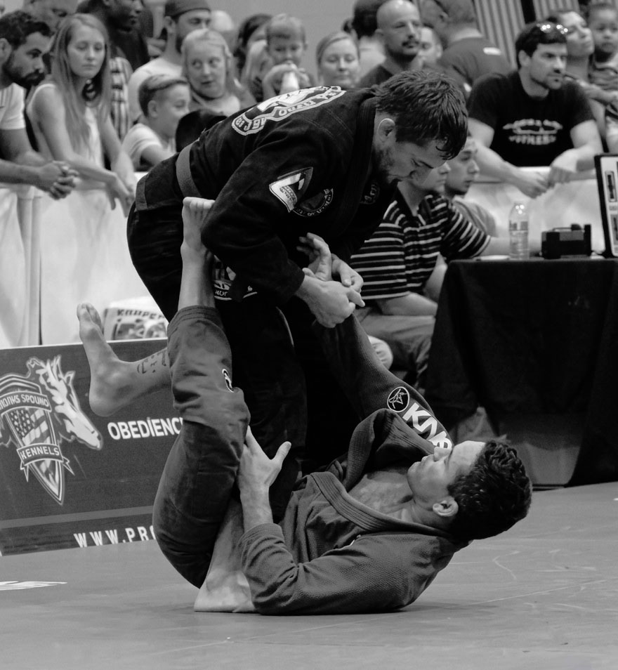 A black belt competitor controlling an opponent's leg during a Jiu Jitsu match
